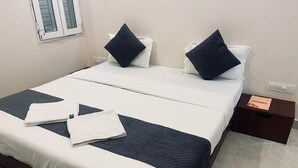 A neatly made bed with white and dark blue bedding, a small bedside table with a book, and a window with closed blinds. - Maa Prabha Courtyard (Varanasi)