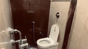 A bathroom with a white toilet, a shower, and a blue bucket. - Maa Prabha Courtyard (Varanasi)