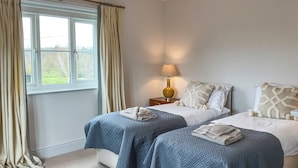 A bedroom with two beds, a window with curtains, a bedside lamp, and a view of greenery outside. - Elm Tree House - UK51762 (Landford, near Salisbury)