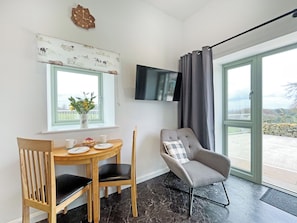 A room with a dining table, chairs, a TV mounted on the wall, and a view through a large window. - Primrose Cottage - UK51766 (Pickering)