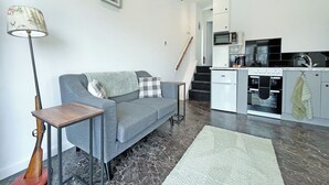 Ein modernes Wohnzimmer mit grauem Sofa, einem kleinen Tisch, einer Stehlampe und einer Küchenzeile mit Mikrowelle, Backofen und Spüle.