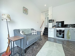 A modern living room with a grey sofa, a small table, a floor lamp, and a kitchen area with a microwave, oven, and sink. - Primrose Cottage - UK51766 (Pickering)