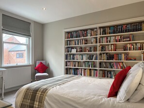Quarto com uma grande estante, janela com vista para um edifício de tijolos, radiador e um travesseiro vermelho em uma cadeira branca.