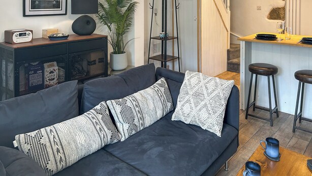 A modern living room with a dark sofa, a coffee table, a plant, a radio, and a staircase leading to a kitchen area. - 2 bedroom accommodation in St Agnes (St Agnes)