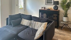 A modern living room with a dark grey sofa, two patterned throw pillows, a wooden coffee table with two black mugs, a small side table with a radio and a plant, and a framed picture on the wall. - 2 bedroom accommodation in St Agnes (St Agnes)