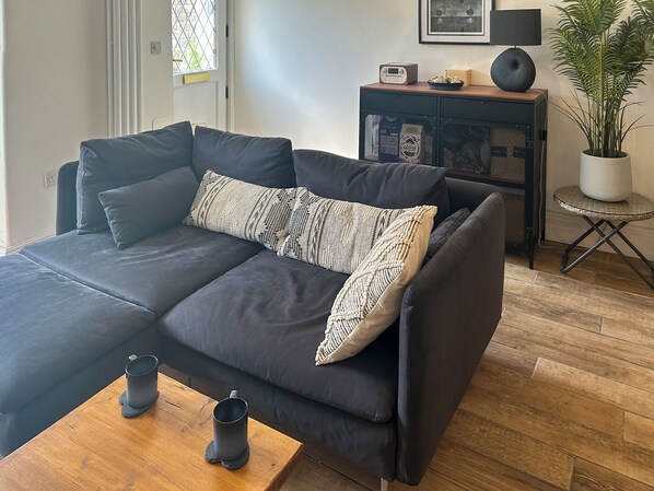 Una sala moderna con un sofá gris oscuro, dos almohadones estampados, una mesa de centro de madera con dos tazas negras, una mesita auxiliar con una radio y una planta, y un cuadro enmarcado en la pared.