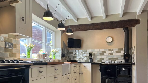 Une cuisine avec des meubles blancs, une cuisinière noire, une horloge au mur et une fenêtre ornée de fleurs.