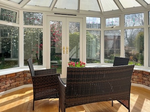 Uma estufa com mobiliário de vime e uma mesa com flores, grandes janelas e uma parede de tijolos.
