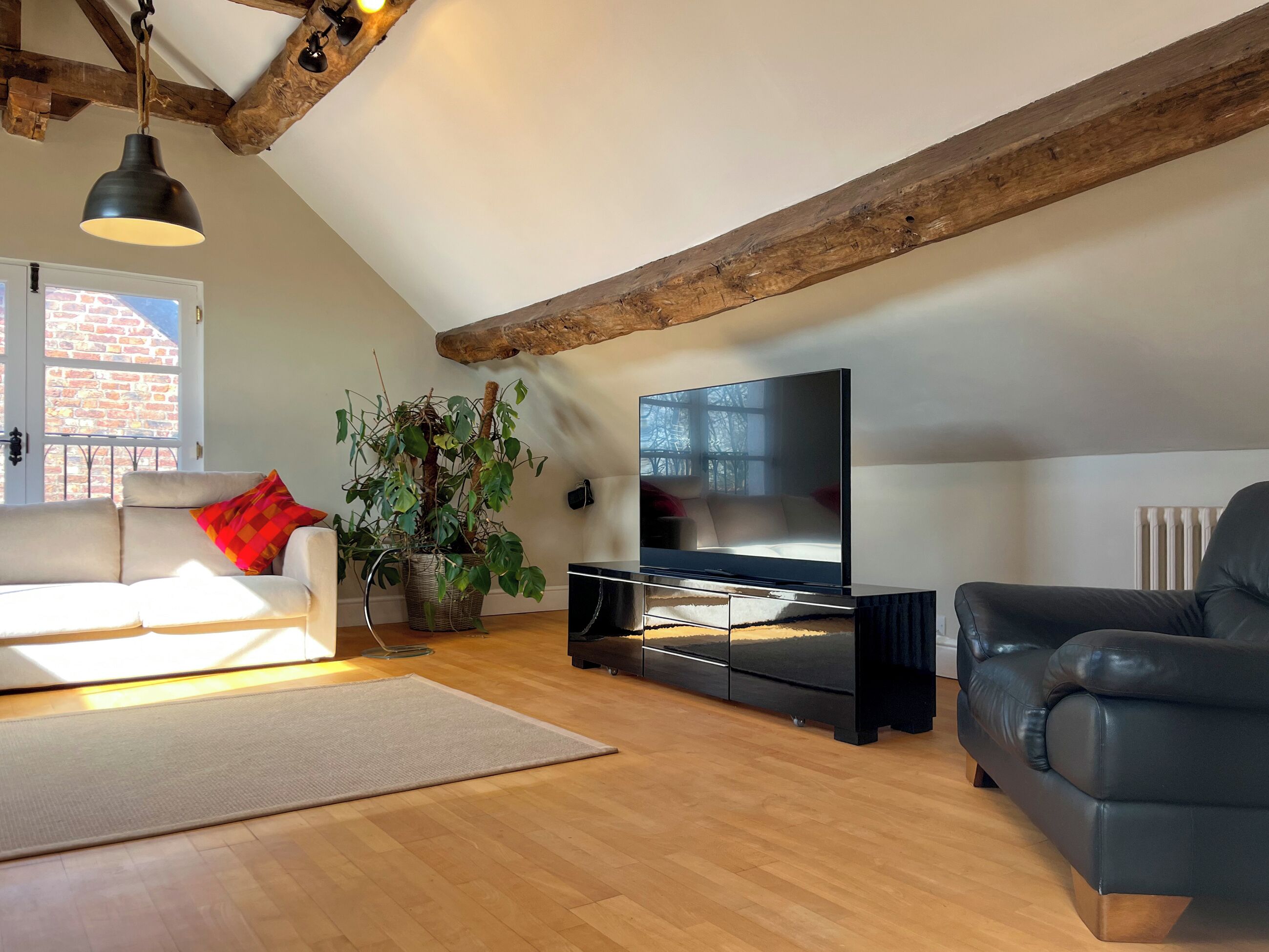 A loft-style living room with exposed wooden beams, a large flat-screen TV, a sofa, a leather armchair, and a potted plant.