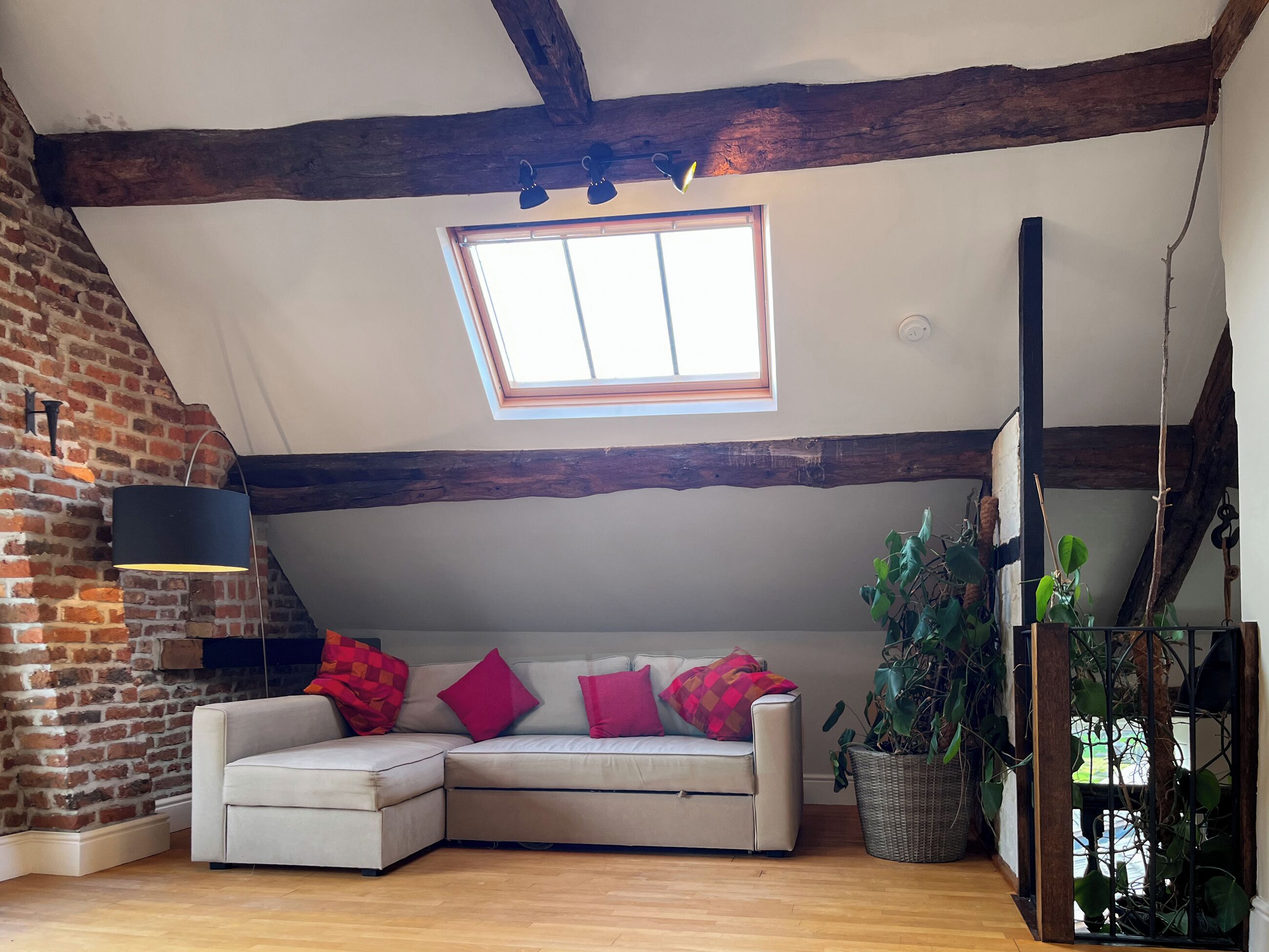 Une pièce de style loft avec des poutres en bois apparentes, un mur en briques, un canapé blanc agrémenté de coussins rouges, une grande fenêtre et une plante en pot.
