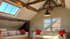 Un espace de vie de style loft comprenant deux canapés, un tapis, des poutres en bois, une lucarne et une fenêtre donnant sur un immeuble en briques.