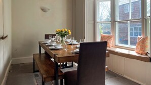 Un coin repas avec une table en bois dressée pour quatre personnes, des bancs, un vase rempli de fleurs jaunes et une fenêtre donnant sur un bâtiment en briques.