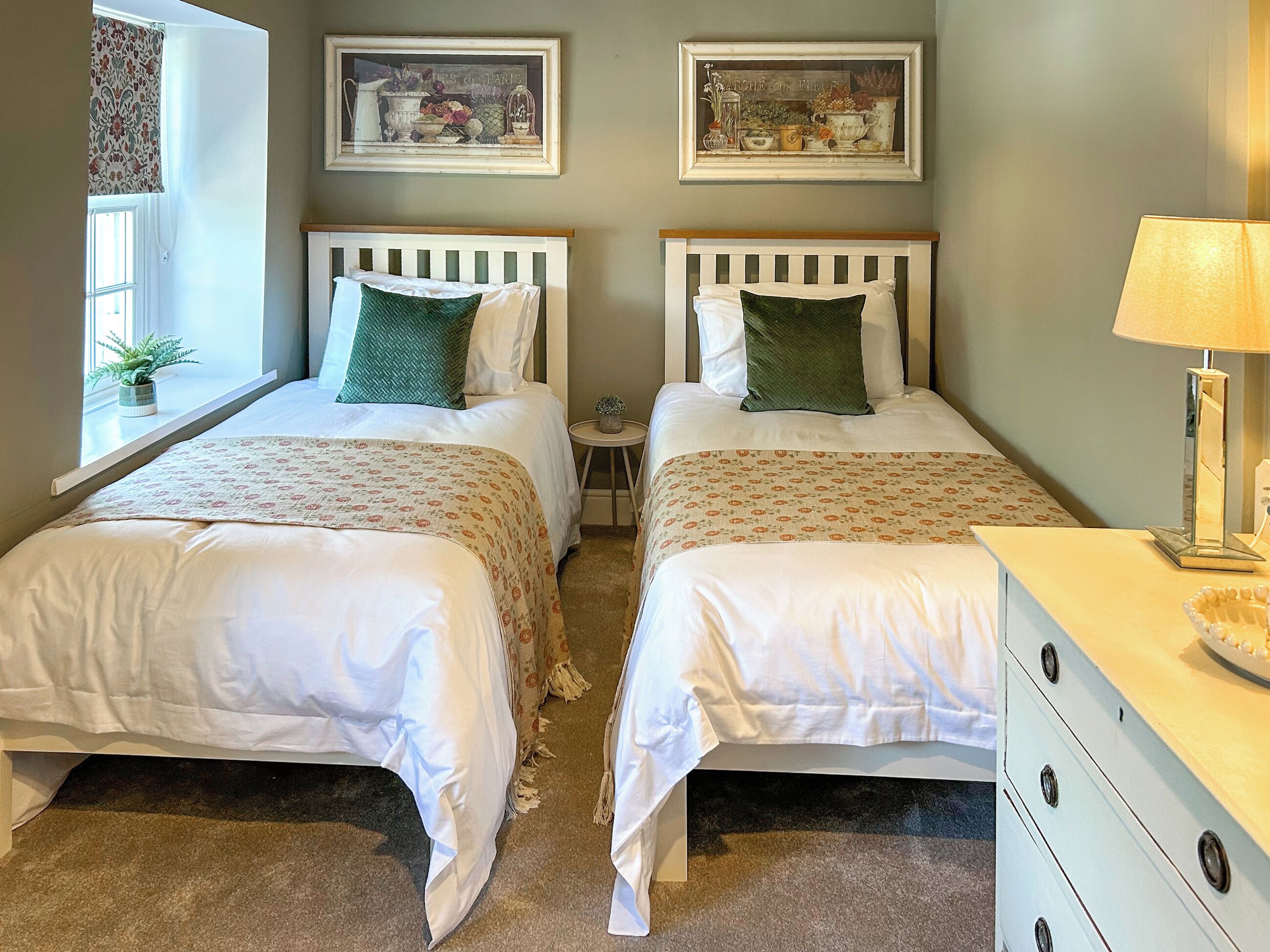 A double bed room with two beds, a small side table, a dresser, and two framed pictures on the wall.