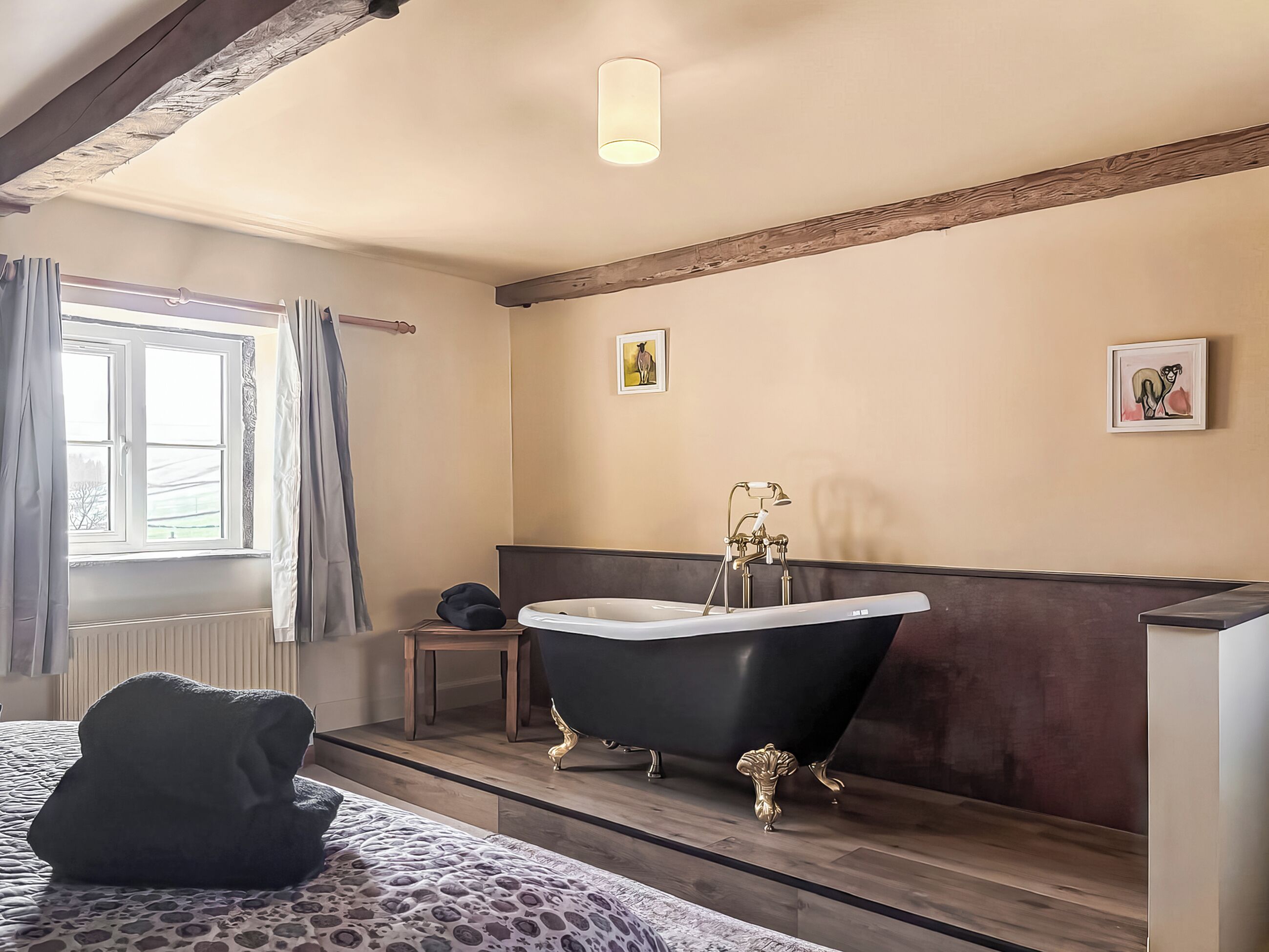 Ein Schlafzimmer mit Bett, einem Fenster mit Vorhängen, einem Heizkörper, einer schwarzen Badewanne mit goldenen Füßen und einem Holzbalken an der Decke.