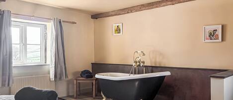 Ein Schlafzimmer mit Bett, einem Fenster mit Vorhängen, einem Heizkörper, einer schwarzen Badewanne mit goldenen Füßen und einem Holzbalken an der Decke.