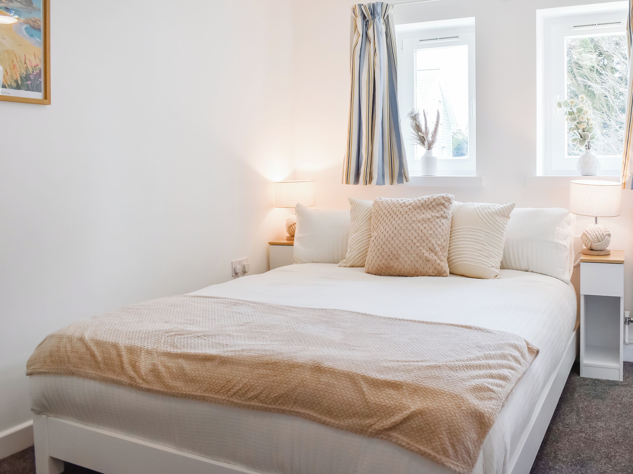 Een slaapkamer met een bed, nachtkastjes, lampen, een raam met gordijnen en een schilderij aan de muur.