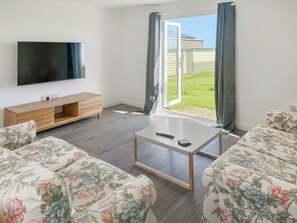 Una sala de estar con televisor, mueble bajo de madera, sofás estampados, una mesa de centro y un ventanal corredizo a un jardín.