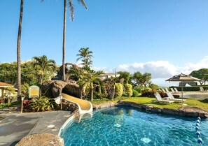 A pool with a slide, lounge chairs, palm trees, and a clear sky. - Beachfront Resort. Ocean Views. (Lihue)