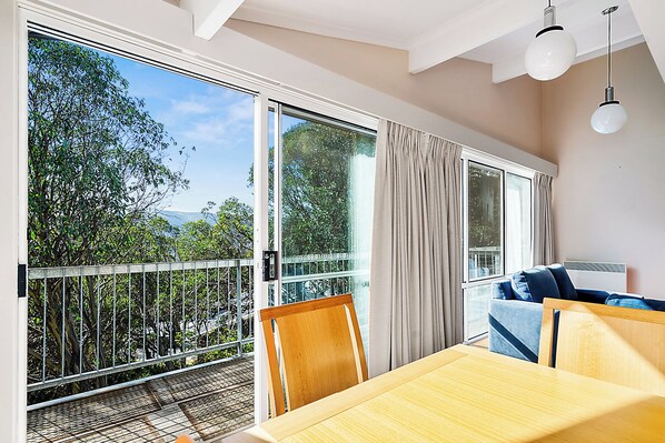 A room with a dining table, chairs, a sofa, and a balcony with a view of trees and a building.