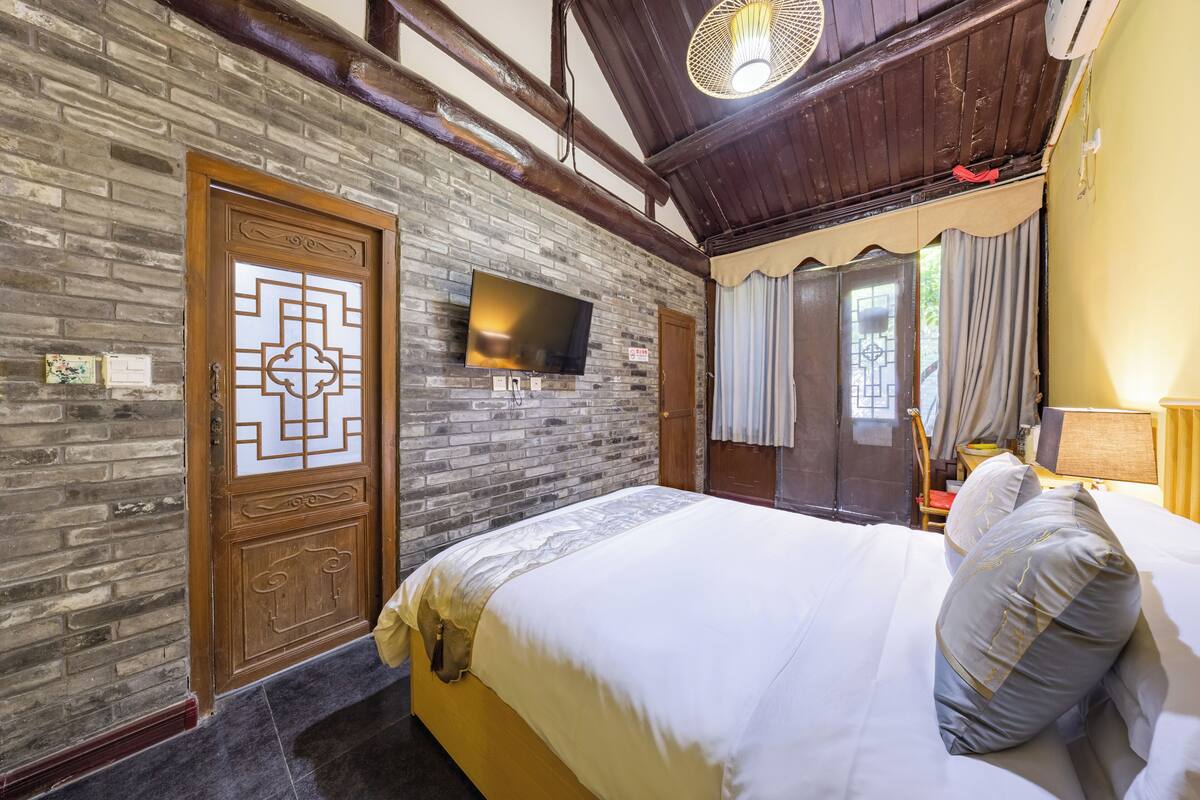 Ein Schlafzimmer mit Ziegelwand, einer Holztür mit Fliegengitter, einem Flachbildfernseher, einem Bett mit weißer Bettwäsche und einer Holzbalkendecke.