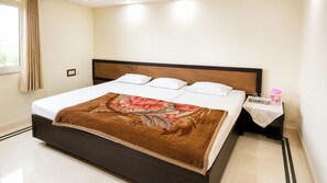 A neatly made bed with a floral bedspread, a wooden headboard, and a nightstand with a small table and a pink container. - Hotel Maharaja Palace AGRA (Agra)