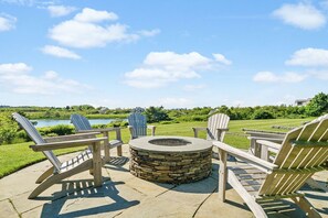 A stone fire pit surrounded by Adirondack chairs in a landscaped outdoor area with a view of a lake and hills.