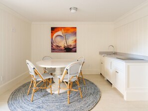 Un comedor con una mesa redonda y cuatro sillas de mimbre, un cuadro de un velero en la pared, y una cocina blanca con un lavamanos.