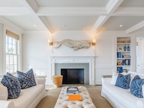 A living room with a fireplace, two sofas with patterned cushions, a coffee table with books, and a bookshelf with decorative items.