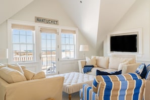 Una habitación con techo alto, un ventanal con vistas, un televisor de pantalla plana montado en la pared y un cartel sobre el ventanal que dice 'NANTUCKET'.