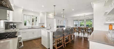 A modern kitchen with white cabinetry, a central island with bar stools, and a dining area with a view of greenery.