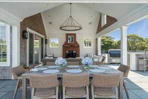 A covered outdoor dining area with a brick fireplace, wicker furniture, and a hanging light fixture.
