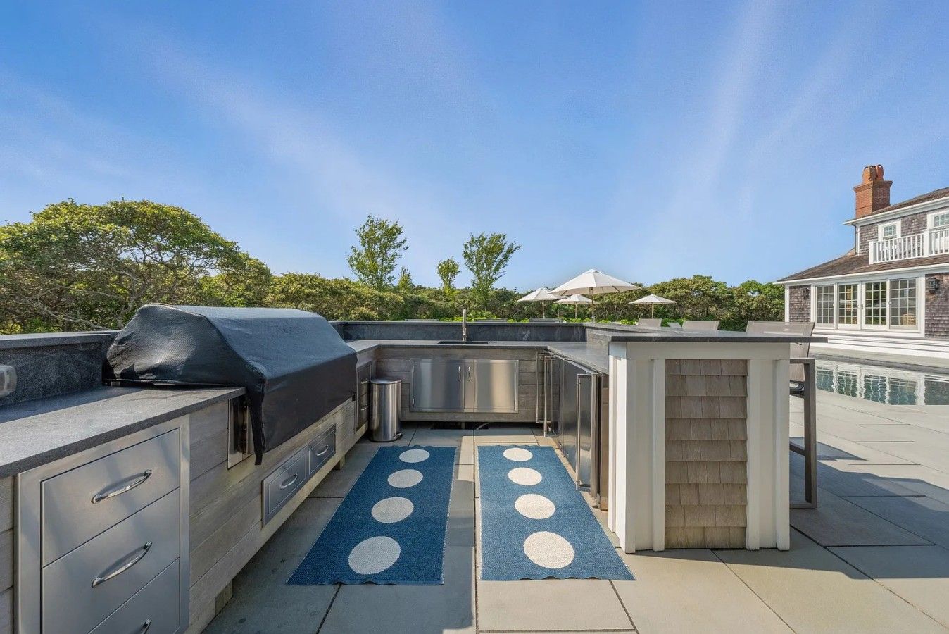 Een buitenkeuken met een barbecue, spoelbak en opbergkasten, uitkomend op een zwembadgedeelte met parasols.
