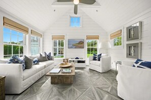 A spacious living room with a high ceiling, white walls, and large windows. There are two white sofas with patterned cushions, a coffee table, and a television.