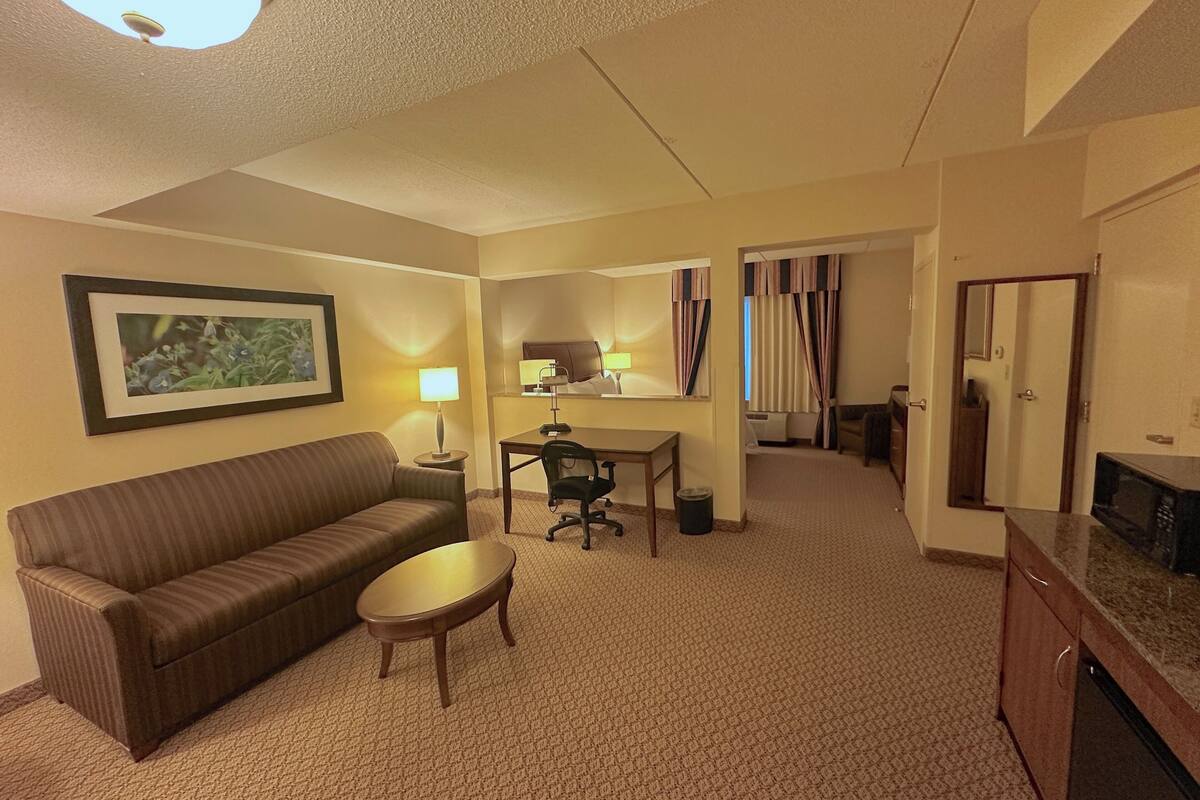 A hotel room with a sofa, a small table, a desk with a chair, a microwave, and a door leading to another room.