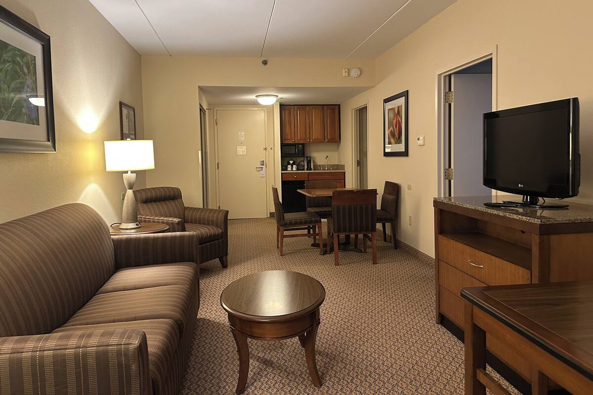 A hotel room with a sofa, a small table, a TV, a kitchenette, and a dining area.