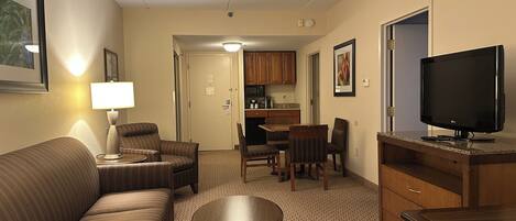 A hotel room with a sofa, a small table, a TV, a kitchenette, and a dining area.