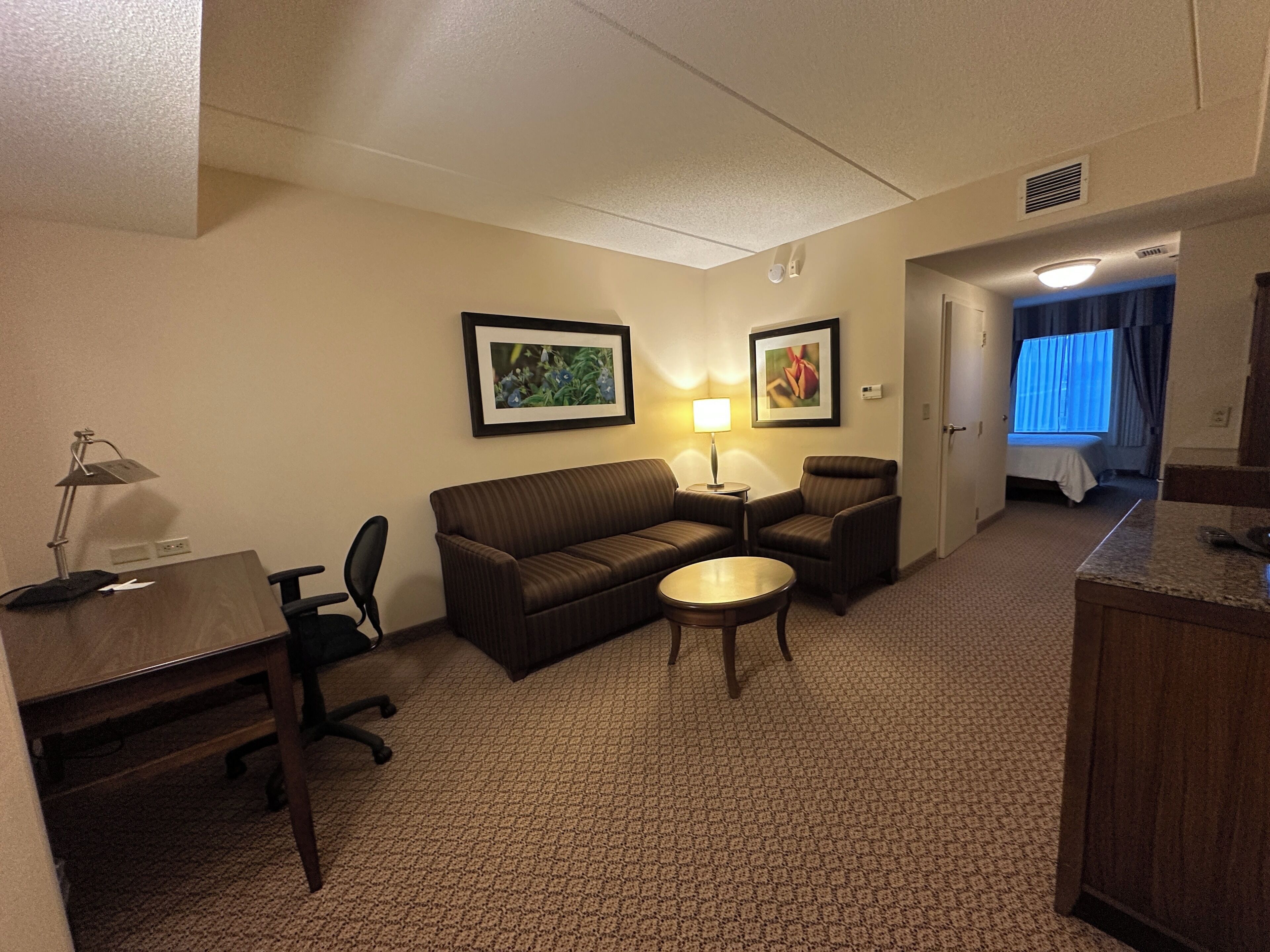 A hotel room with a sofa, two armchairs, a small table, a desk with a lamp, and a bed in the background.