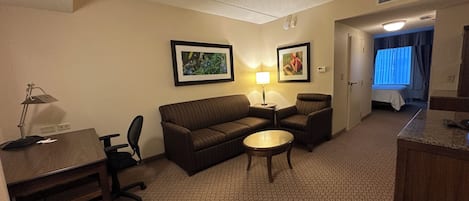 A hotel room with a sofa, two armchairs, a small table, a desk with a lamp, and a bed in the background.