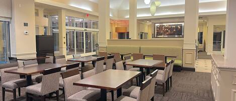 A hotel dining area with multiple tables and chairs, a television, and a kitchen area in the background.