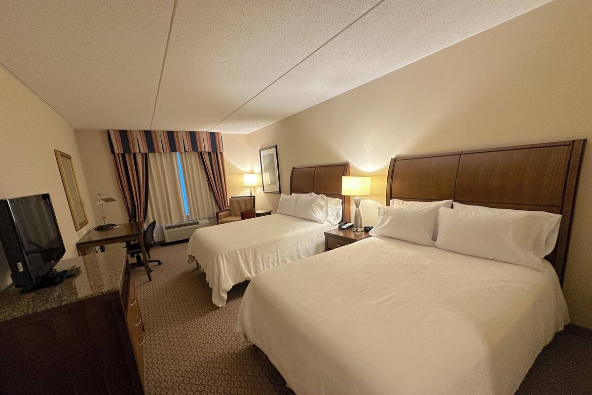 A hotel room with two beds, a desk, a television, and a window with curtains.