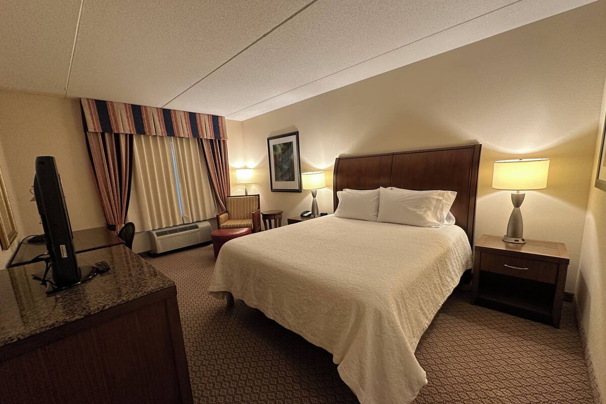 A hotel room with a bed, a desk with a computer, a chair, a nightstand with a lamp, and a wall-mounted picture.
