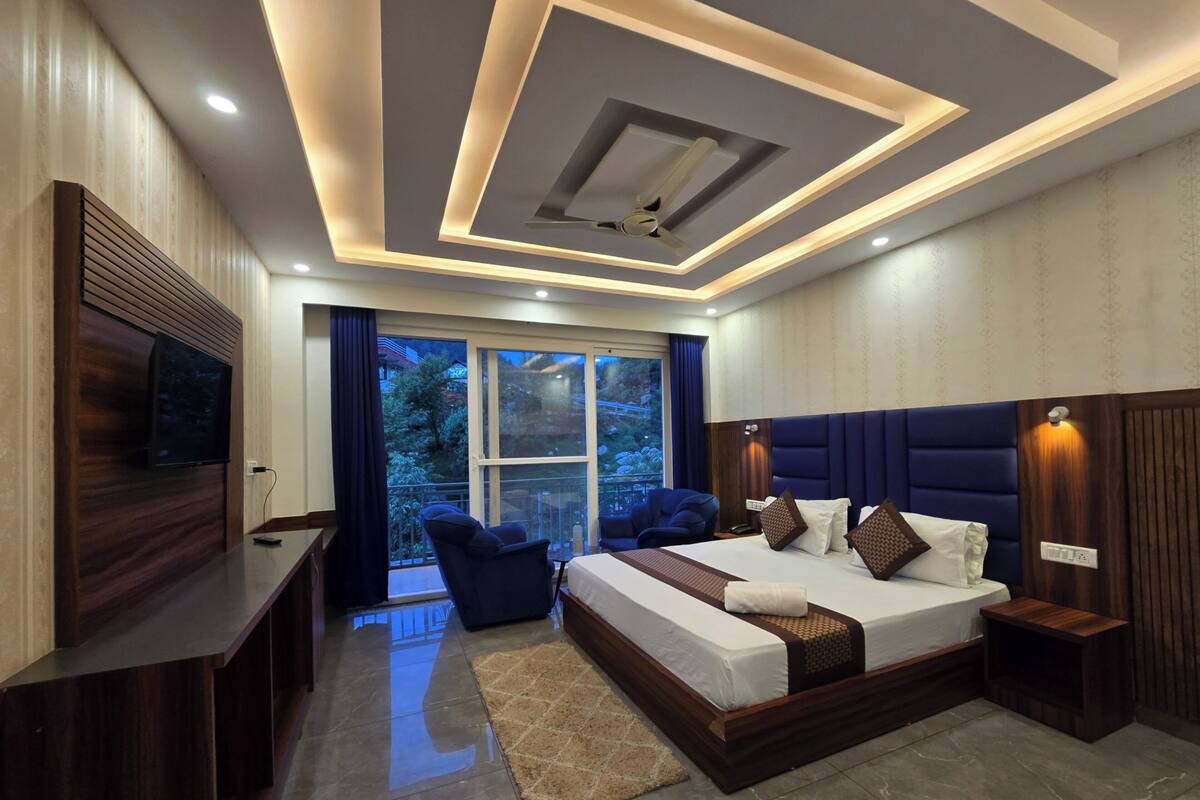 A modern bedroom with a large bed, a TV, a balcony with a view, and a ceiling with recessed lighting.