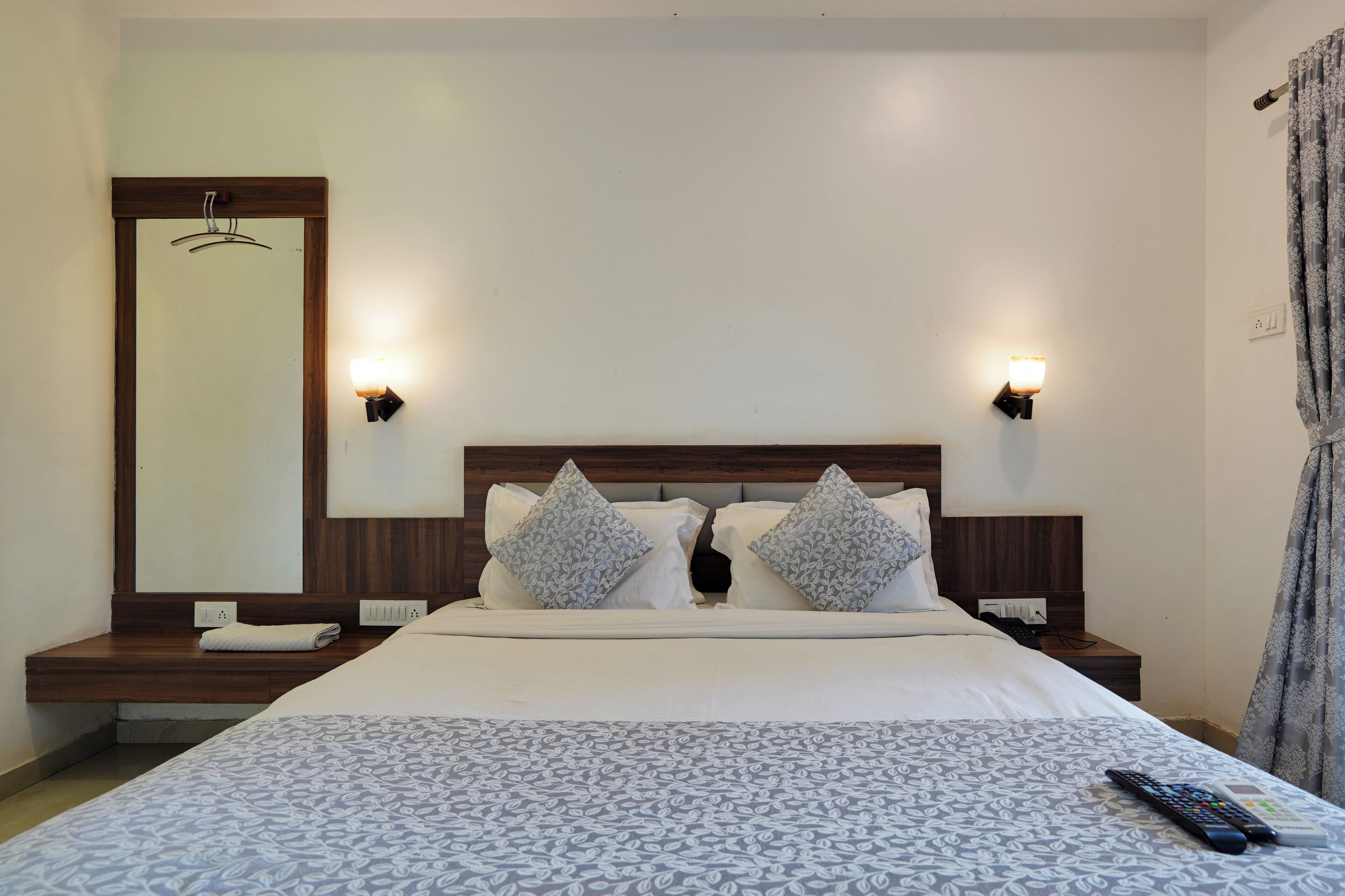 A hotel room with a bed, two bedside tables, a nightstand, a mirror, and a curtain.