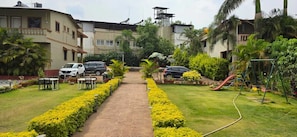 A residential area with a paved pathway, parked cars, a playground, and a building with a balcony. - Prince Palace Bhilar in Village of Books (Bhilar)