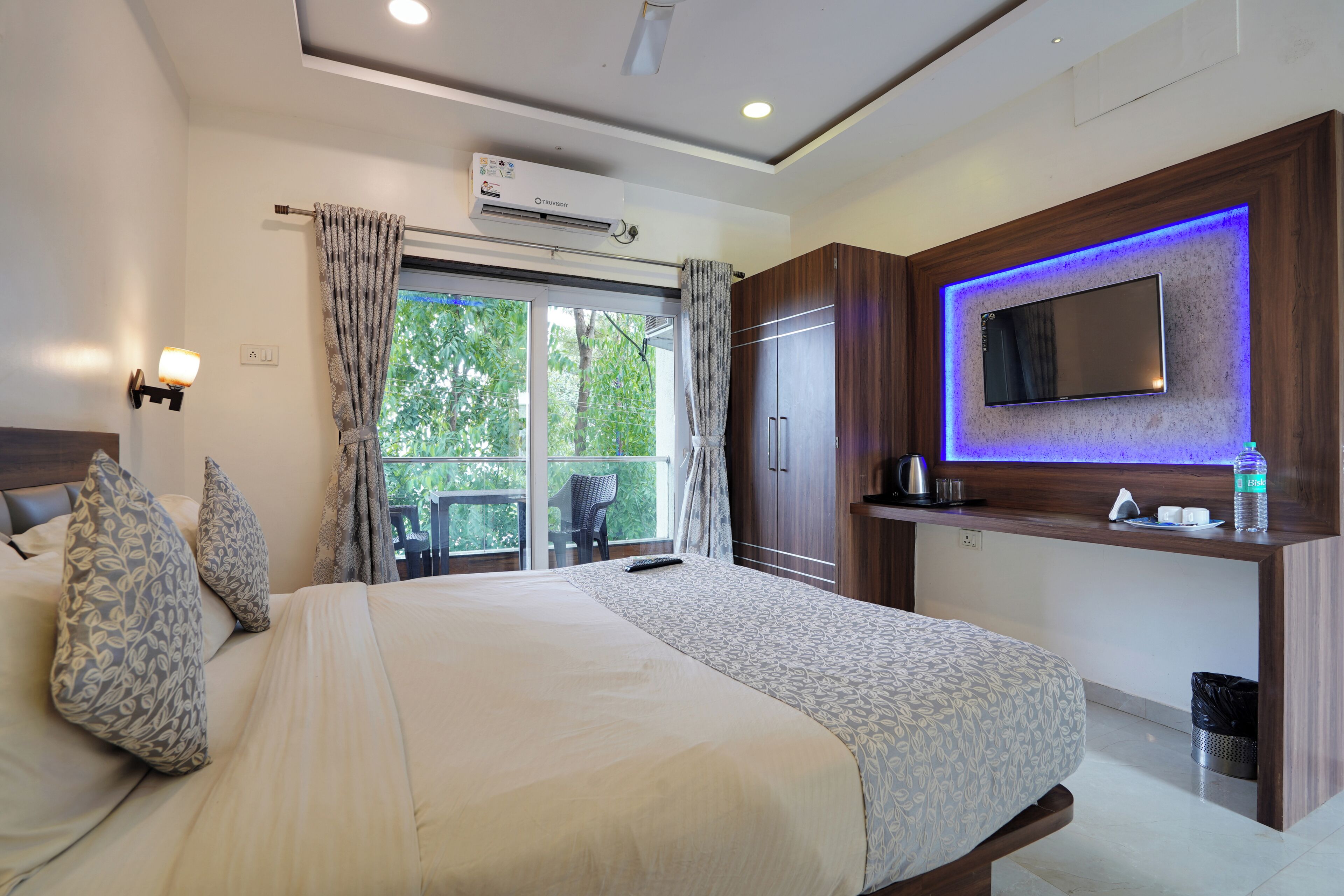 Habitación de hotel con cama, televisor montado en un panel de madera, ventana con cortinas y unidad de aire acondicionado.