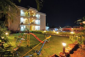 A multi-story hotel with a well-lit lawn, a children's play area, and a pool in the background.