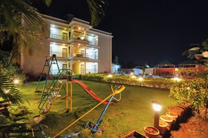 Un hotel de varios pisos con un césped bien iluminado, una zona de juegos para niños y una piscina al fondo.