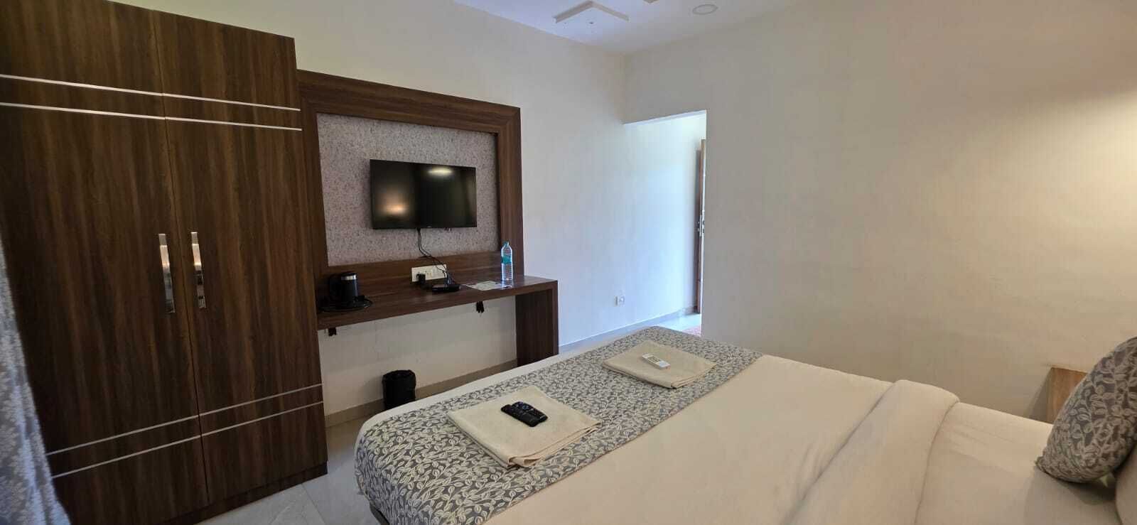 A hotel room with a bed, a TV mounted on a wooden panel, a desk with a chair, and a window.