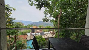 A balcony with a metal railing, a black table, and a chair. In the background, there are trees, a building with a red roof, and a mountainous landscape. - Prince Palace Bhilar in Village of Books (Bhilar)