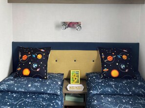 Ein Schlafzimmer mit einem Bett, zwei mit Weltall-Motiven versehenen Kissen, einem kleinen Tisch mit Tablett und einem an der Wand befestigten Regal mit einem Spielzeugauto.